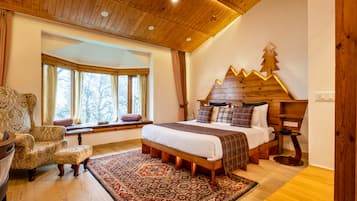 Ein Schlafzimmer mit Holzboden, einem Bett mit Holz Kopfteil, einem Fenster mit Vorhängen, einem gemusterten Sessel und einem kleinen Tisch mit Lampe.