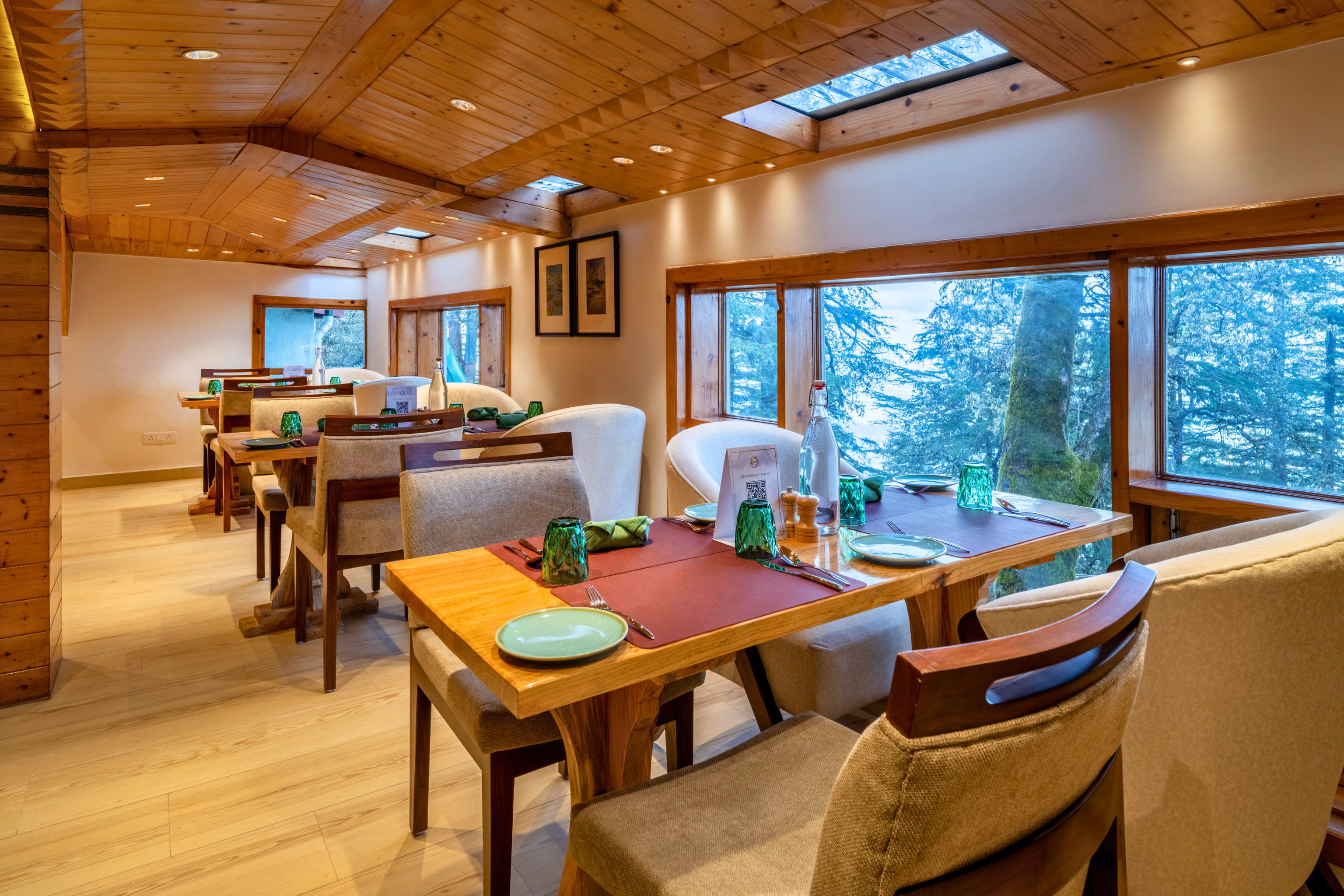 A dining area with wooden tables and chairs, large windows with a forest view, and a high ceiling with wooden beams.