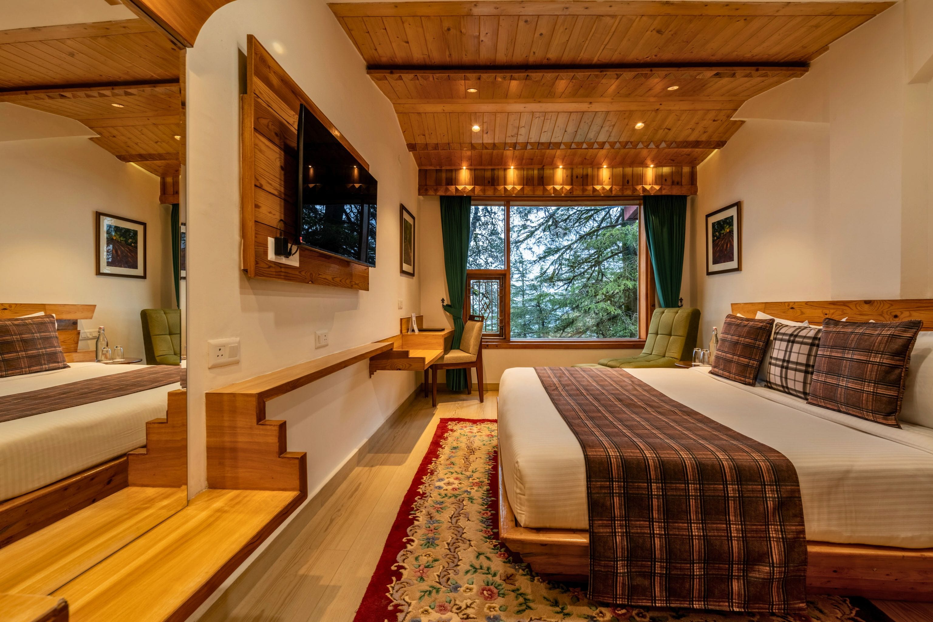A hotel room with two beds, a TV, a desk, and a window with a view of trees.