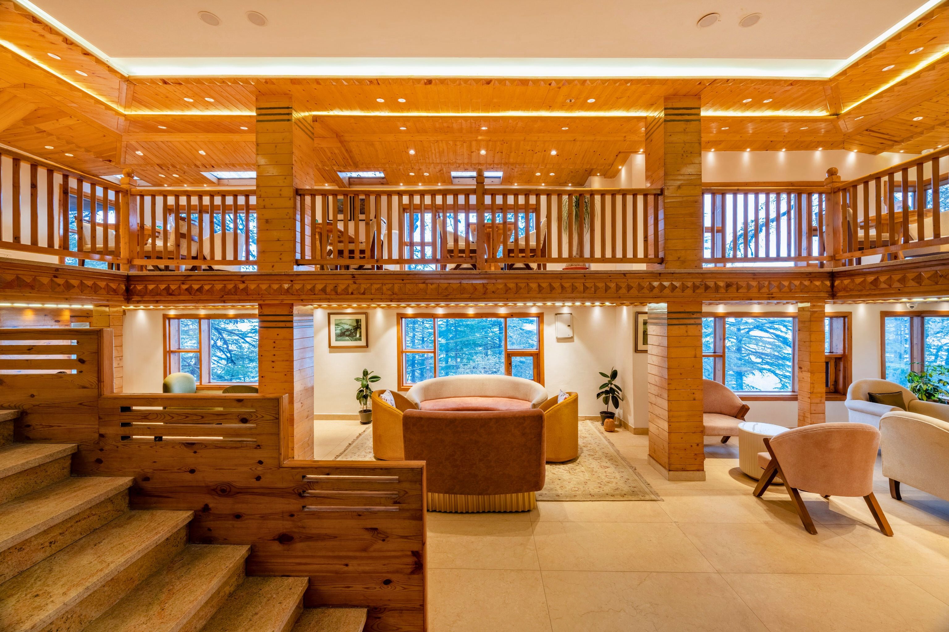 A spacious interior with a wooden staircase leading to a mezzanine level, featuring a seating area with sofas and a view of the outdoors.