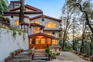 A two-story house with a prominent front entrance, a staircase leading to the main door, and a surrounding landscape with trees.