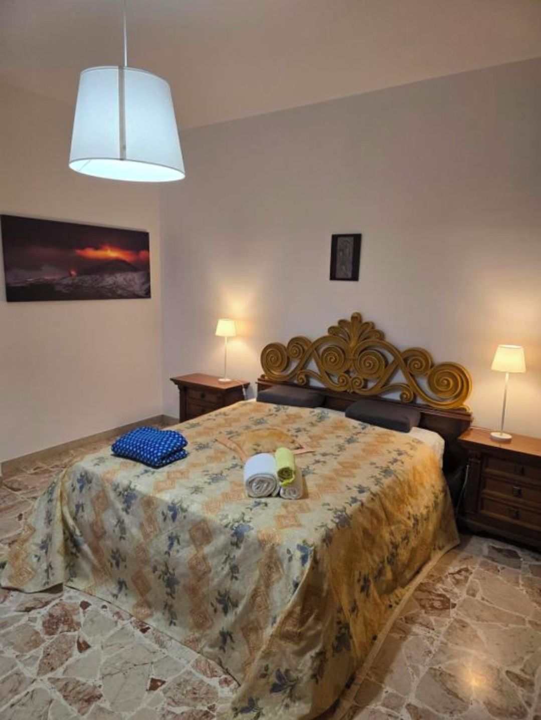 A bedroom with a bed, two bedside tables, a lamp, a painting, and a headboard.