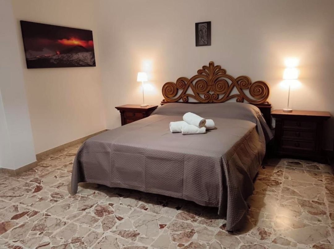 A bedroom with a stone floor, a bed with a brown bedspread, two bedside lamps, and a painting on the wall.