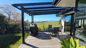 A covered outdoor patio with a table and chairs, a barbecue grill, and a view of a green field and mountains. - Mapua View! Surrounded by rolling pasture and manicured gardens (Richmond)