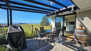 A covered outdoor area with a table and chairs, a barbecue grill, and a view of a green landscape. - Mapua View! Surrounded by rolling pasture and manicured gardens (Richmond)