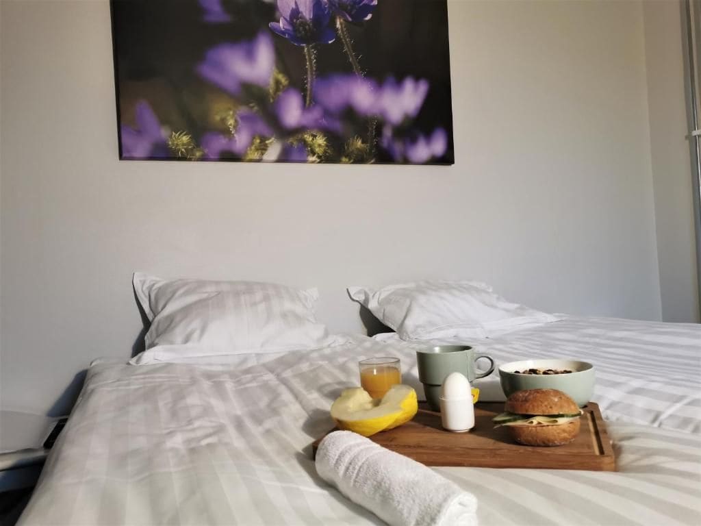 Un lit bien fait, avec du linge de lit blanc, un panier de fruits contenant une banane et une pomme, une tasse, un bol de céréales et une serviette enroulée sur un plateau en bois.