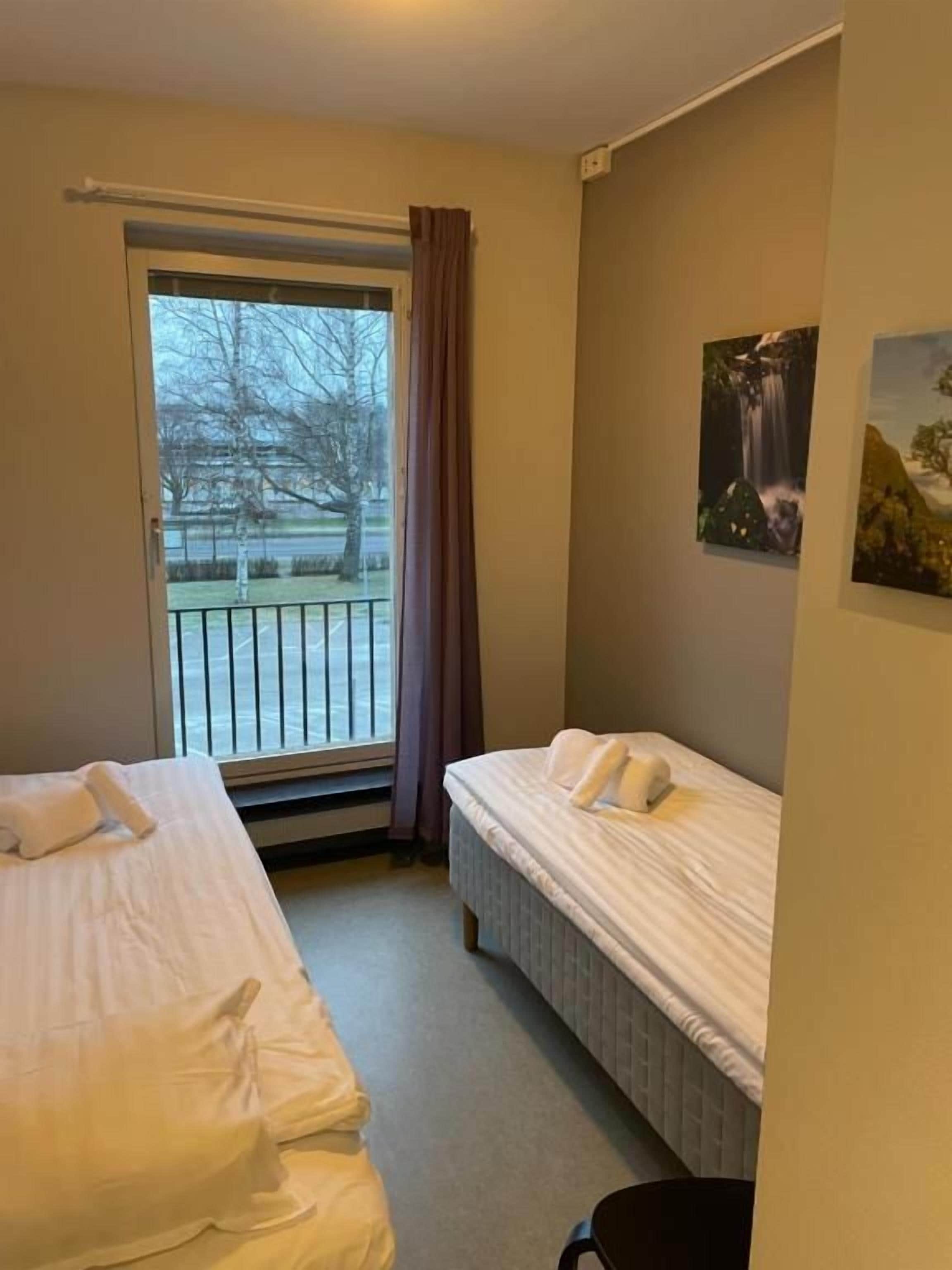 A hotel room with two beds, a window with a view of a park, and a painting on the wall.