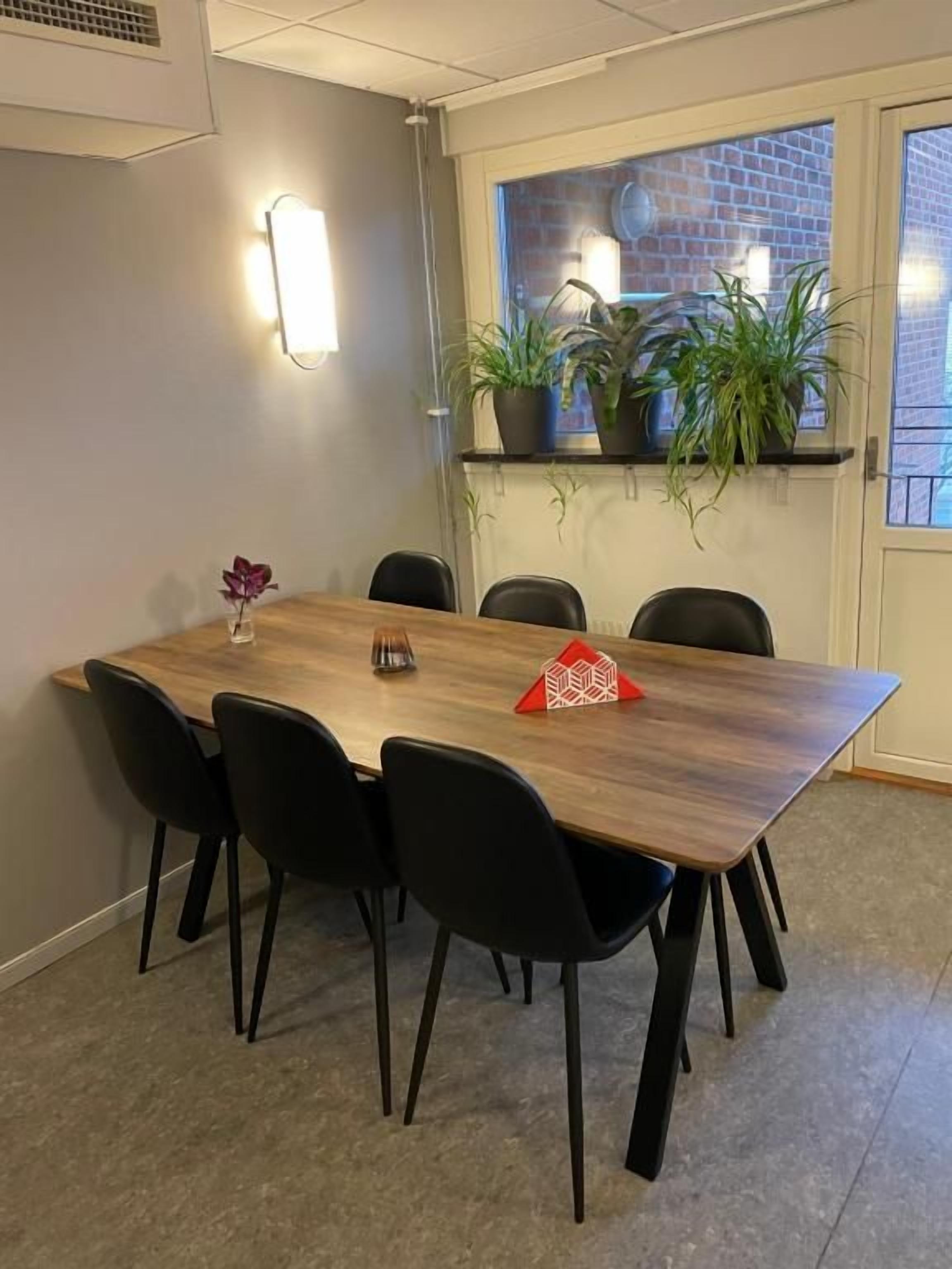 Une salle de conférence avec une table en bois, des chaises noires, une plante en pot et une fenêtre donnant sur un bâtiment en briques.