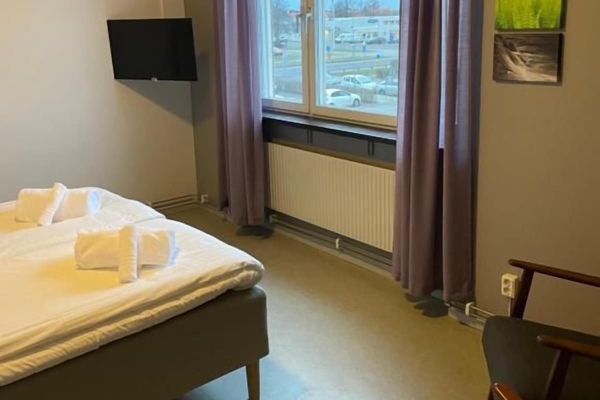 Una habitación de hotel con cama, una silla, televisor, ventana con cortinas y un radiador.