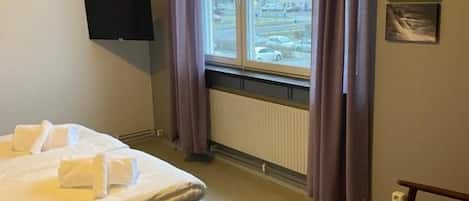 A hotel room with a bed, a chair, a television, a window with curtains, and a radiator.