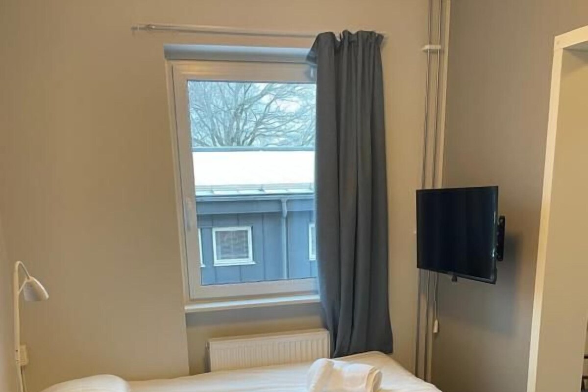 Habitación de hotel con una cama individual, una mesita de noche, una silla, un televisor montado en la pared y una ventana con cortinas.