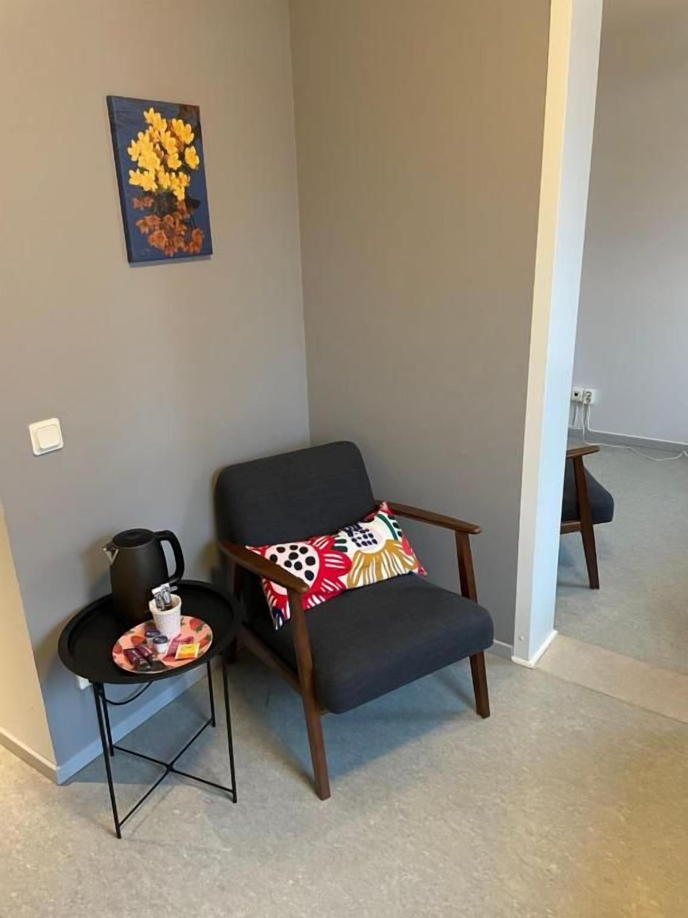 A corner room with a dark-colored armchair, a small table with a kettle, and a wall-mounted artwork.