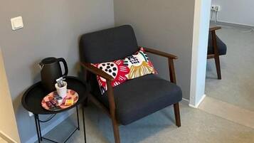 A corner room with a dark-colored armchair, a small table with a kettle, and a wall-mounted artwork.