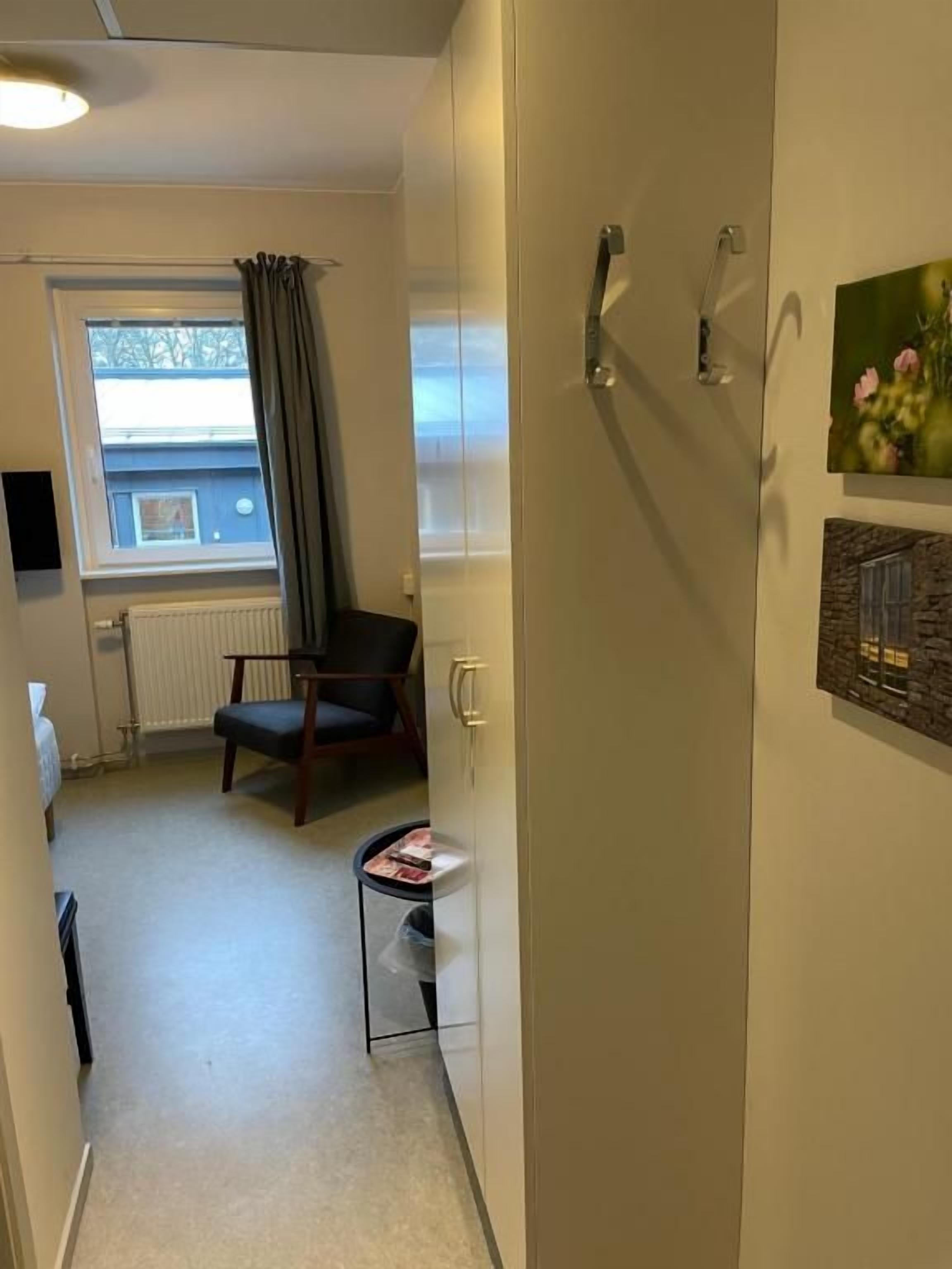 A narrow hallway with a radiator, a chair, a small table with a magazine, and a window with curtains.