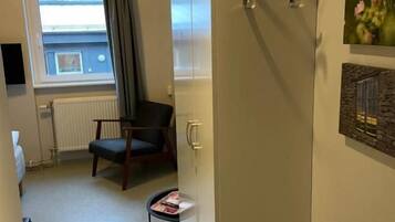 A narrow hallway with a radiator, a chair, a small table with a magazine, and a window with curtains.