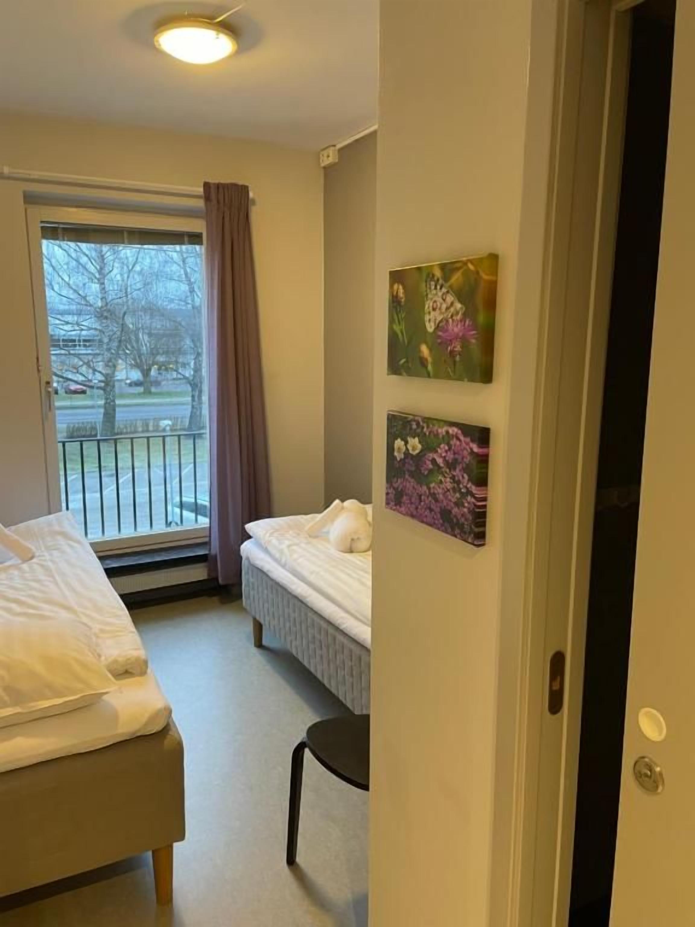 A hotel room with two beds, a window with a view of a tree and a building, and two wall-mounted pictures.