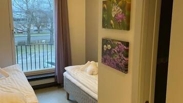 A hotel room with two beds, a window with a view of a tree and a building, and two wall-mounted pictures.