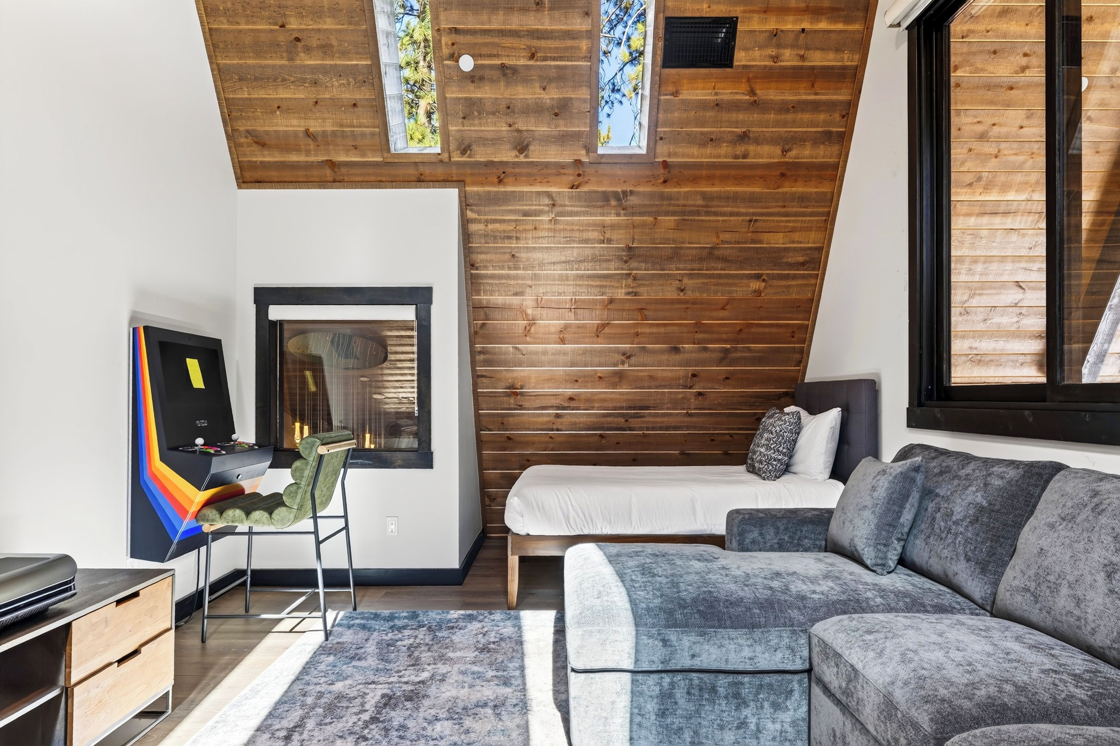 A modern bedroom with a sloped ceiling, wooden paneling, a bed, a grey sofa, and a desk with a colorful chair.