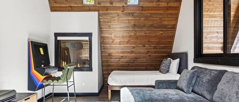 A modern bedroom with a sloped ceiling, wooden paneling, a bed, a grey sofa, and a desk with a colorful chair.