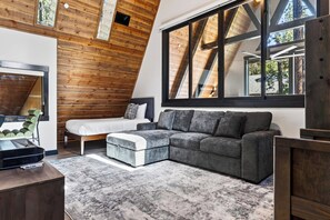 Ein modernes Schlafzimmer mit schräg versiegelter Decke, einem großen Fenster, einem Bett, einem grauen Sofa und einem Schreibtisch mit einem Computer.