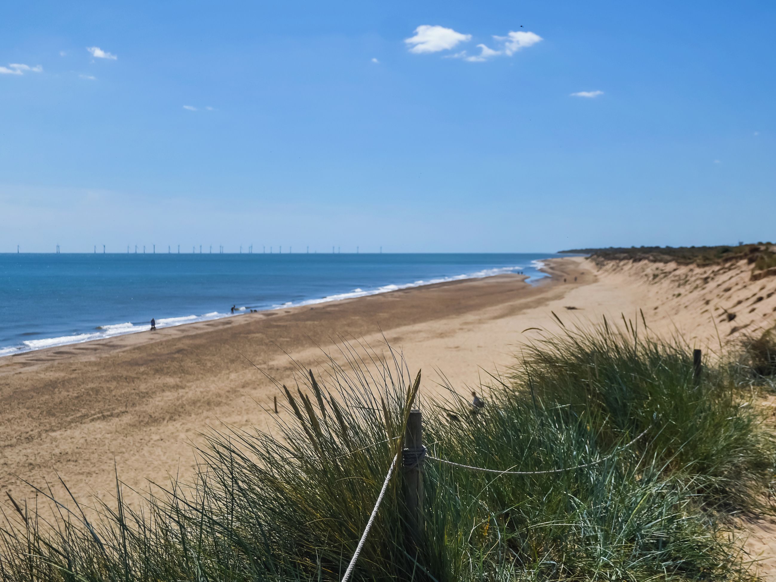Ein Strand mit einem Zaun im Vordergrund, einem klaren Himmel und entfernten Windkraftanlagen.