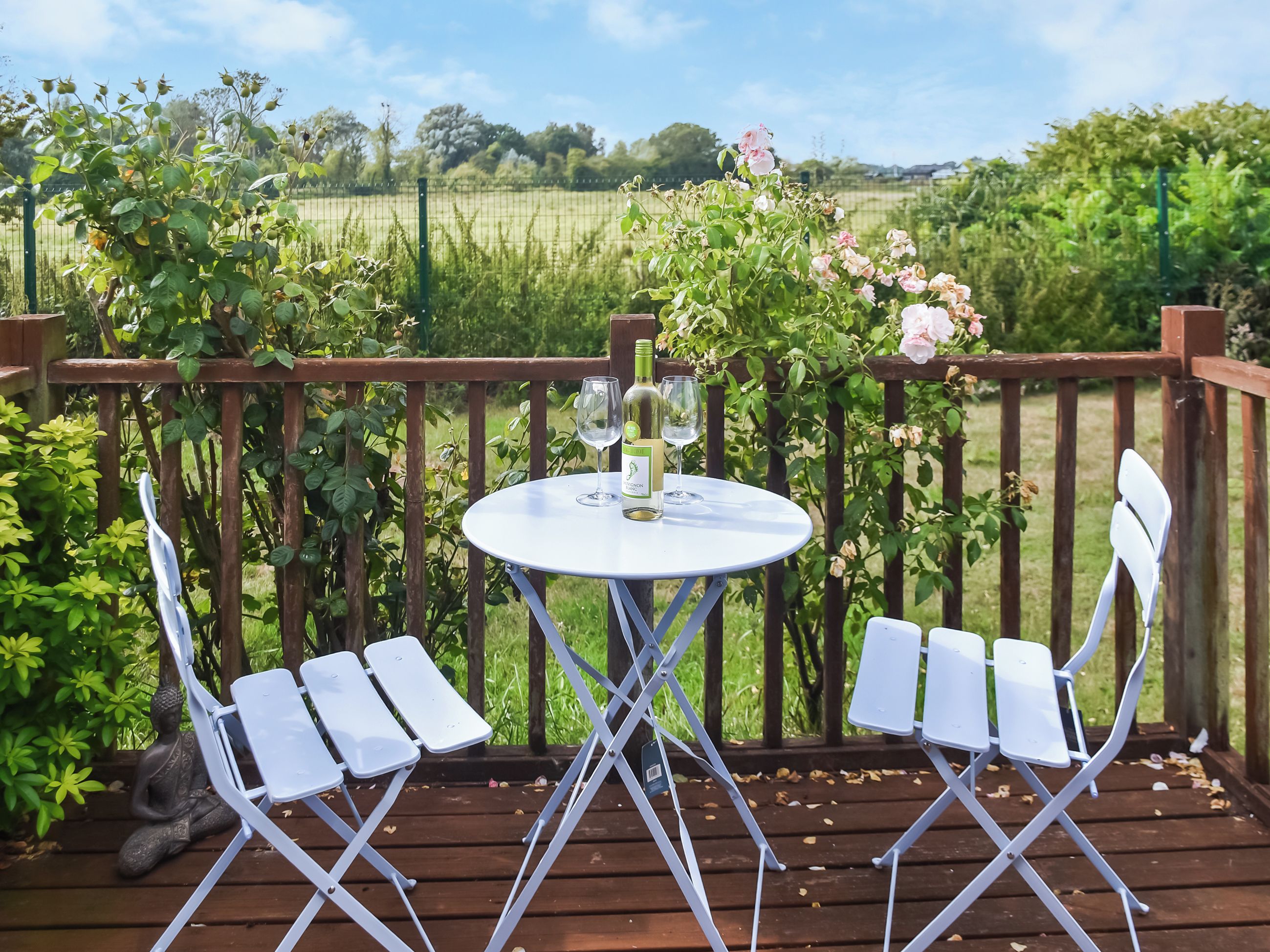 A wooden deck with a white table and chairs, surrounded by greenery and a fence.