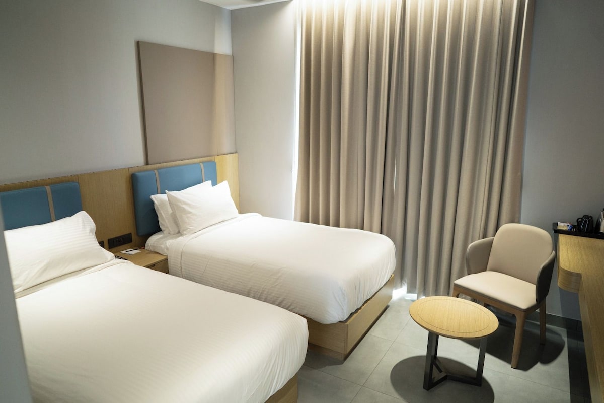 A hotel room with two beds, a chair, a small table, and a curtain.