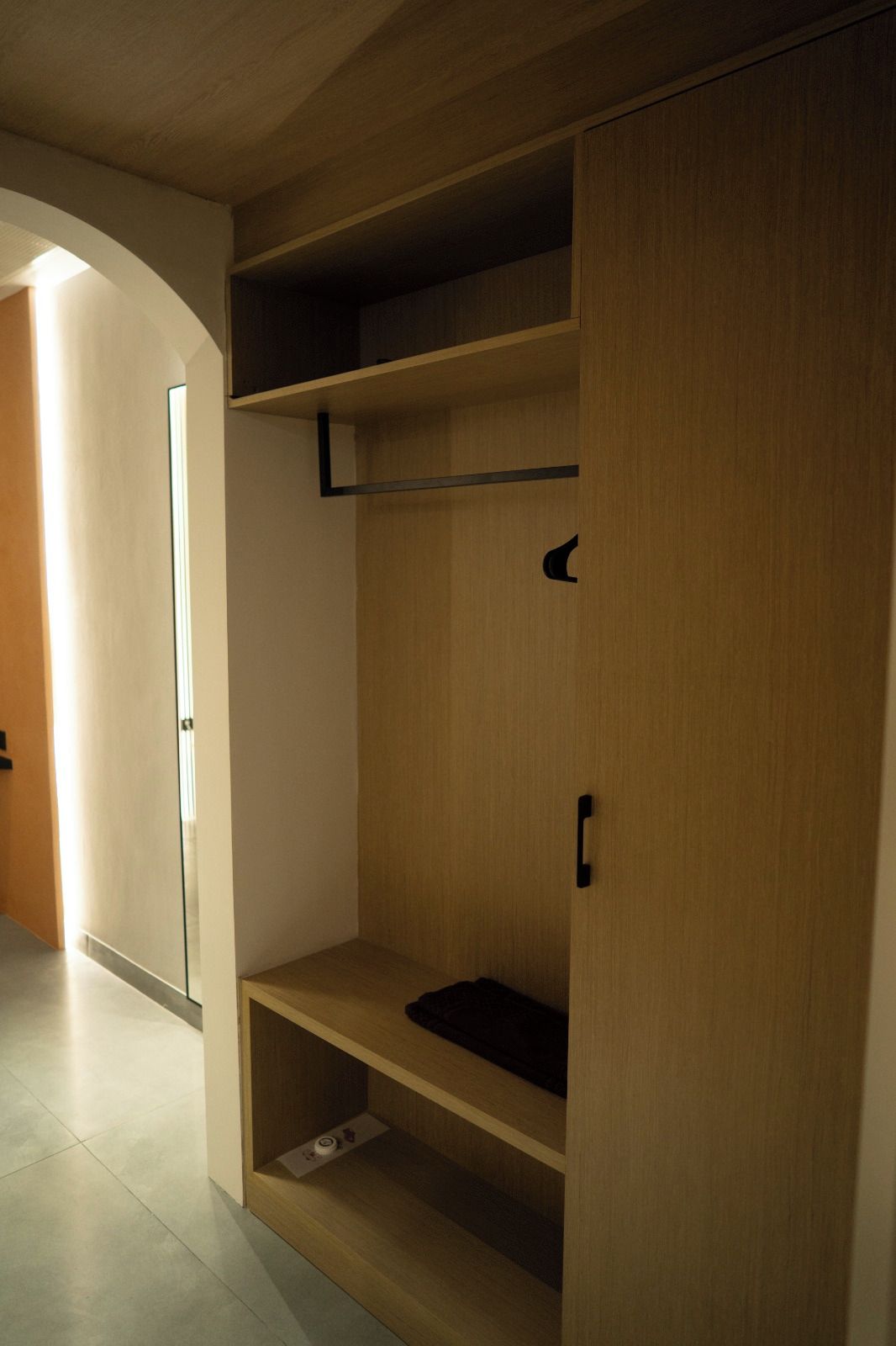 A wooden wardrobe with multiple shelves and a hanging rod, located in a hallway with a glass door on the left.
