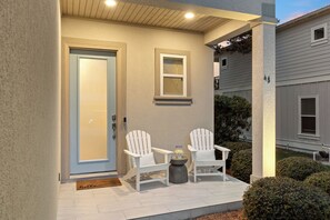 Fachada de casa com porta de entrada, duas cadeiras Adirondack brancas, uma pequena mesa com um jarro e x ícaras, e uma caixa de correio.