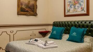 A hotel room with a bed, green upholstered headboard, two teal pillows, a framed floral artwork above the bed, a painting on the wall, and a wooden floor.