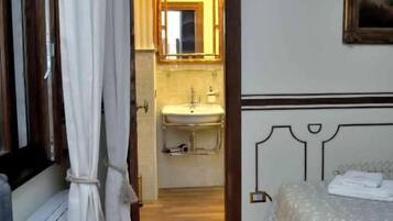 A hotel room with a wooden floor, a bed with a white bedspread, a bedside table, a window with curtains, and a bathroom visible through an open door.
