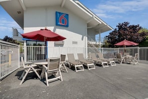 A covered outdoor seating area with lounge chairs and umbrellas, a building with a blue sign displaying the number 6, and a white fence surrounding the space. - Sunny Queen Bed Room|Pool|Free Wifi (Bellingham)