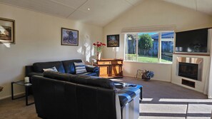 A living room with a black leather sofa, a wooden side table, a fireplace, and a large window with a view of a backyard. - Holiday Haven, a real family getaway (Richmond)