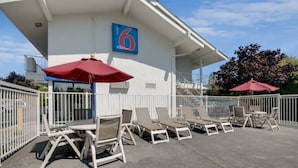 A covered outdoor seating area with lounge chairs and umbrellas, a building with a blue sign displaying the number 6, and a white fence surrounding the space. - Spacious 2-bed Room|Pool|Free Wifi (Bellingham)