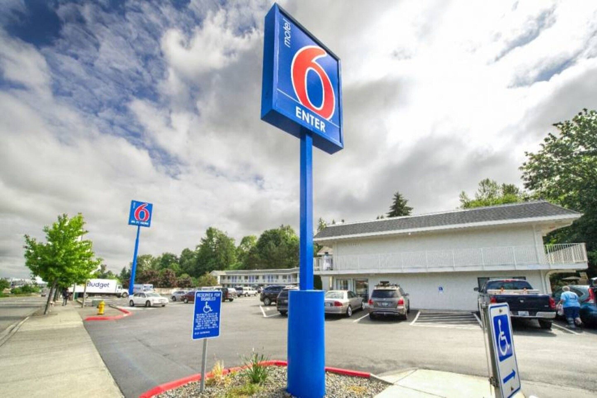 A motel with a blue sign indicating the entrance, a parking lot with several cars, and a building with a white facade.