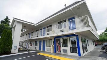 Un motel de dos pisos con un letrero con el número '6', un balcón en el segundo piso y un estacionamiento al frente.