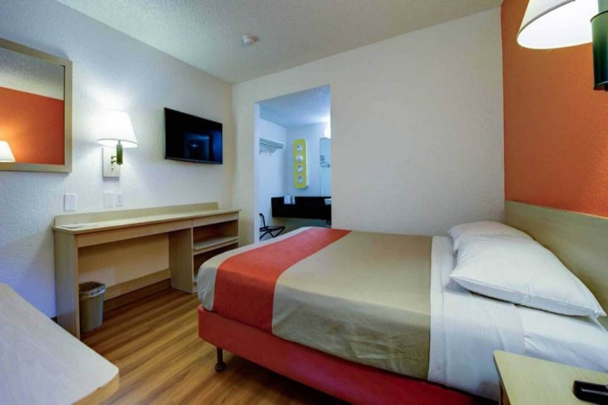 A hotel room with a bed, a desk, a chair, a TV, and a bathroom visible through an open door.
