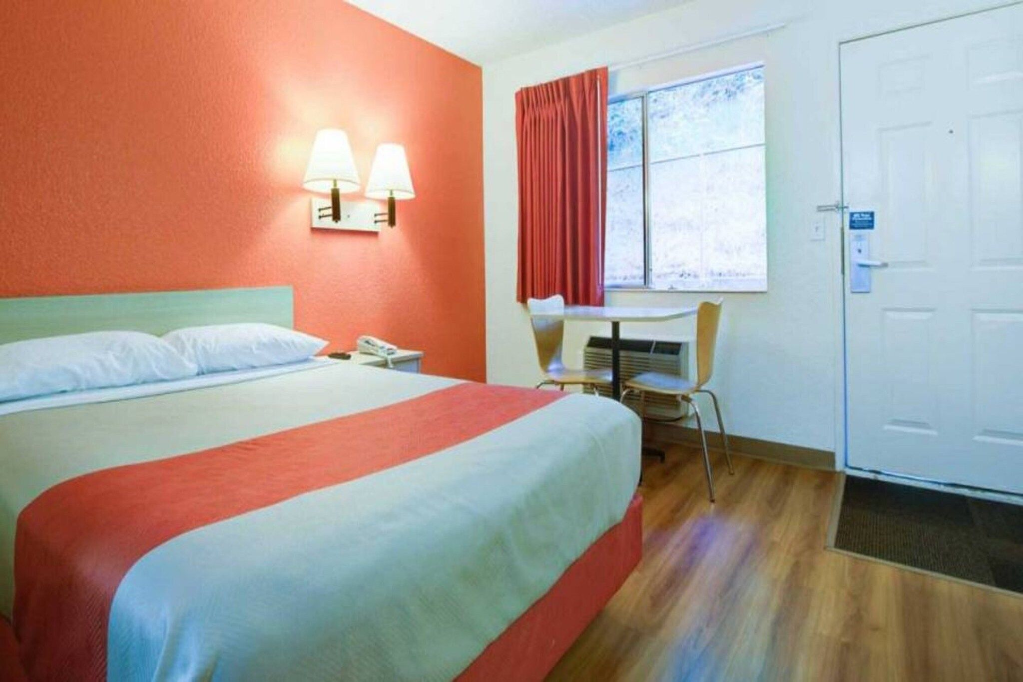 A hotel room with a bed, a desk, a chair, a window with curtains, and a door.