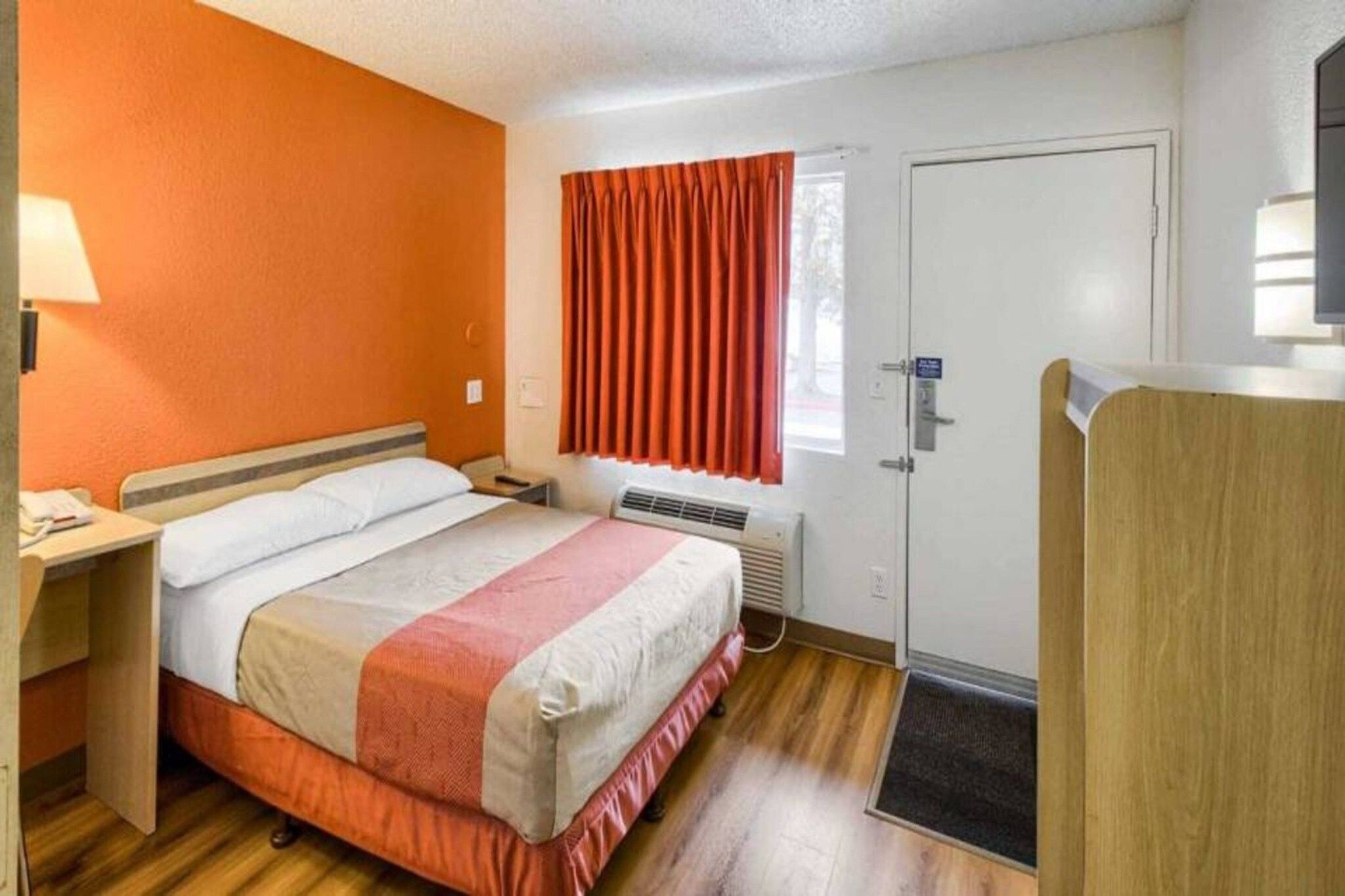 A hotel room with a bed, bedside table, window with curtains, door, and a small desk with a lamp.