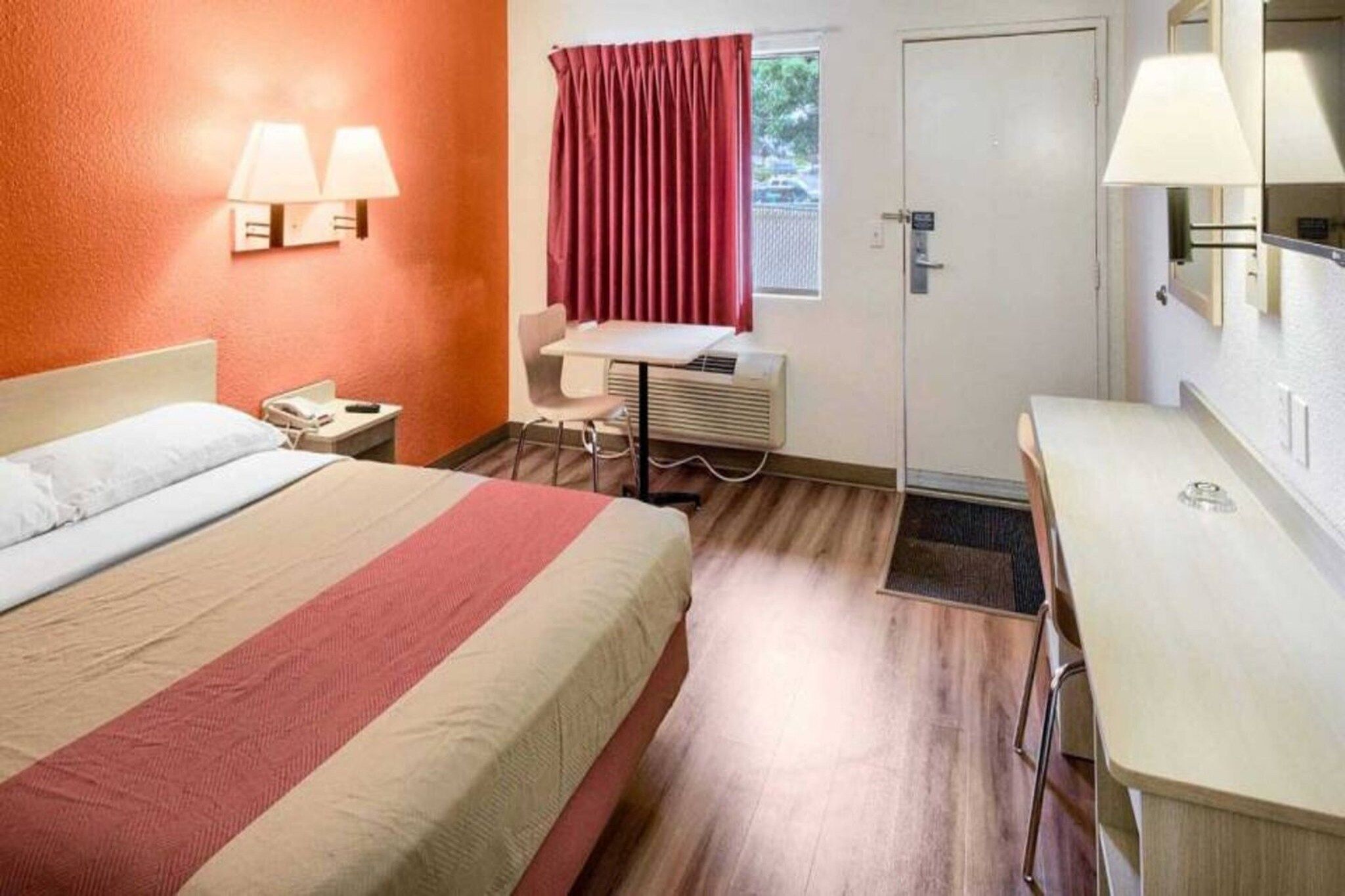 A hotel room with a bed, bedside table, desk, chair, and an air conditioning unit.