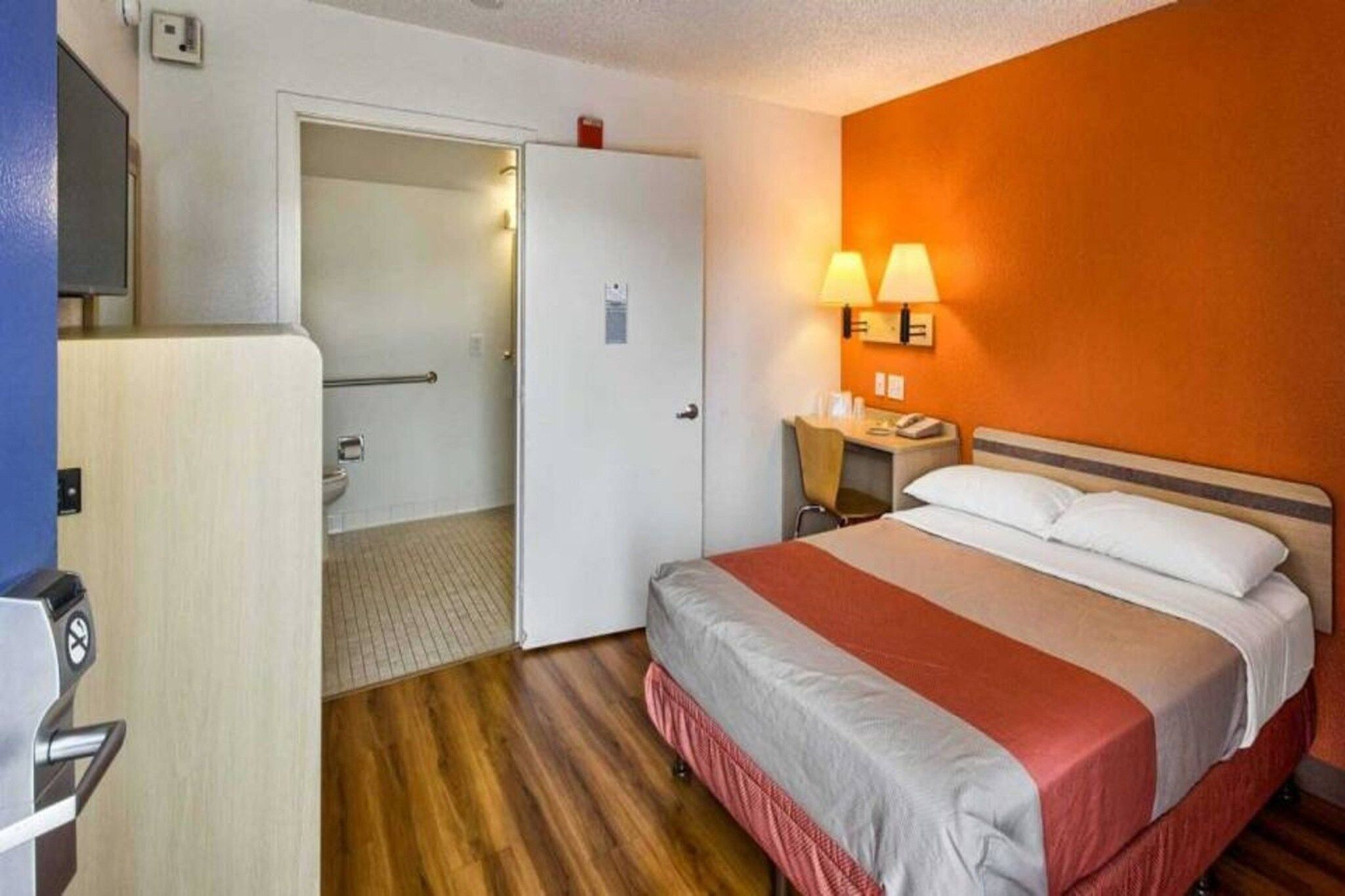 A hotel room with a bed, bedside table, lamp, and a door leading to a bathroom.