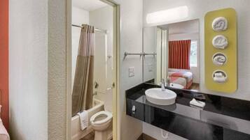 A hotel bathroom with a toilet, sink, and shower. There is a mirror with a towel rack and a bed visible in the background.