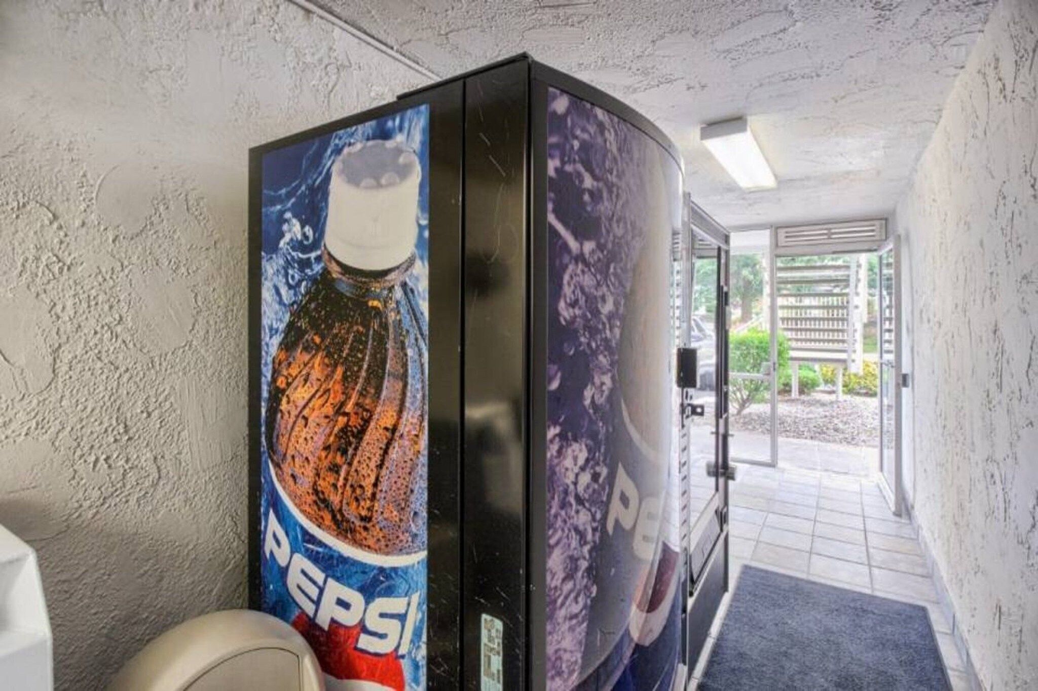 Baño con máquina expendedora de Pepsi, inodoro y puerta que da a un espacio exterior.