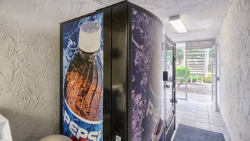 Baño con máquina expendedora de Pepsi, inodoro y puerta que da a un espacio exterior.