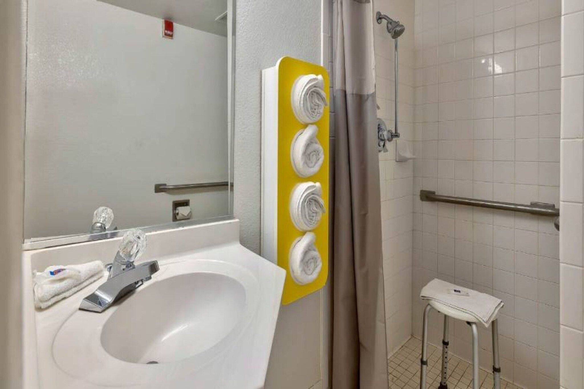 Un baño con un lavamanos blanco, un espejo, un dispensador de toallas amarillo, una ducha con barra de apoyo y un taburete blanco pequeño.