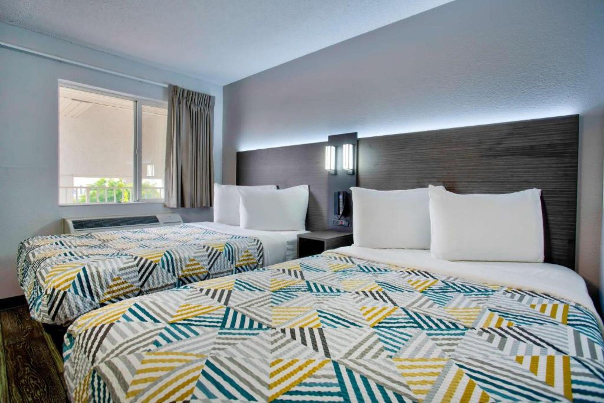 A hotel room with two beds, a window with curtains, a bedside table with a lamp, and a wall-mounted phone.