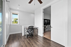 A modern, well-lit room with a wooden floor, a desk with a chair, a window, and a door leading to another room. - Ocean View Lanai + Covered Alfresco Dining | Near Poipu Beach | Hale Pōmaikaʻi (Koloa)