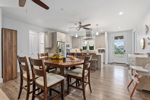 A modern kitchen with wooden cabinets, a dining table with chairs, and a ceiling fan. - Ocean View Lanai + Covered Alfresco Dining | Near Poipu Beach | Hale Pōmaikaʻi (Koloa)