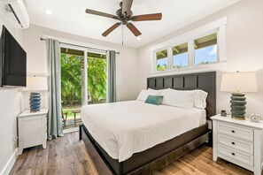 A bedroom with a bed, a ceiling fan, a window with a view of greenery, a bedside table with a lamp, and a nightstand with a clock. - Ocean View Lanai + Covered Alfresco Dining | Near Poipu Beach | Hale Pōmaikaʻi (Koloa)