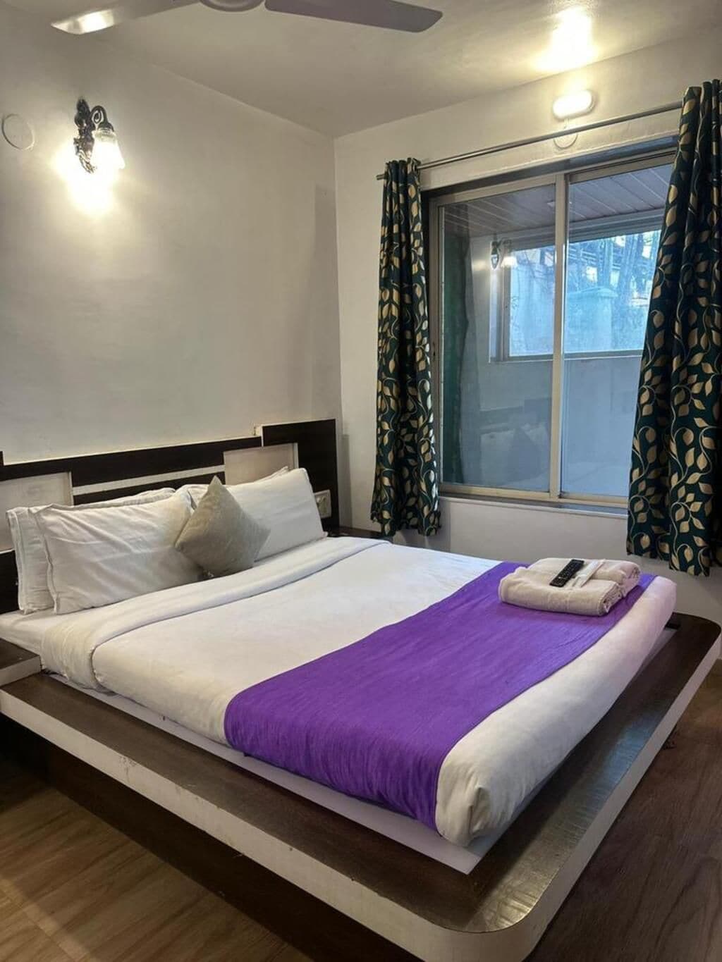 Una camera d'albergo con un letto, una finestra con tende, una ventola a soffitto e un lampadario.